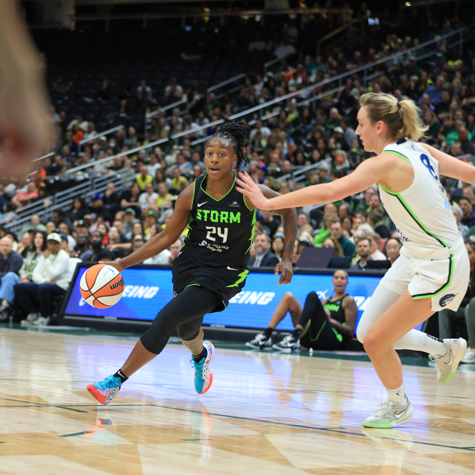 A basketball player in a black jersey dribbles the ball, pursued by a player in a white jersey, during a game with spectators in the background.