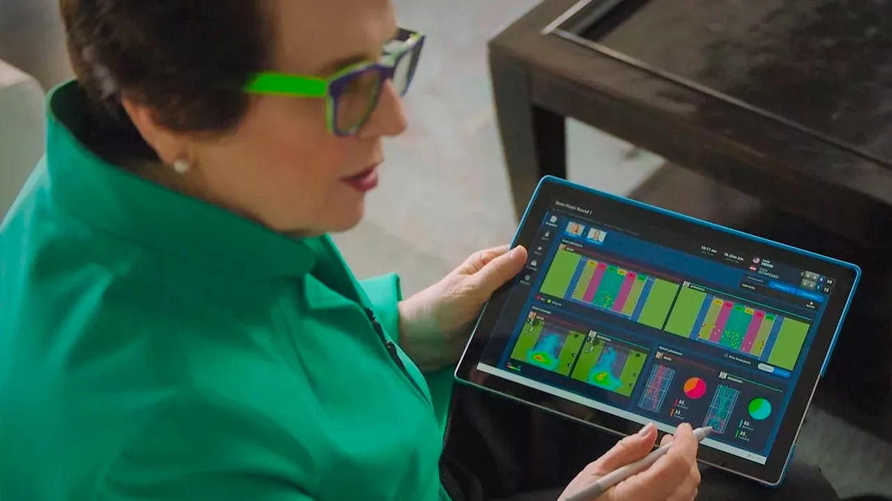 A woman holds a Microsoft Surface with tennis dashboard on the screen while talking to another girl.