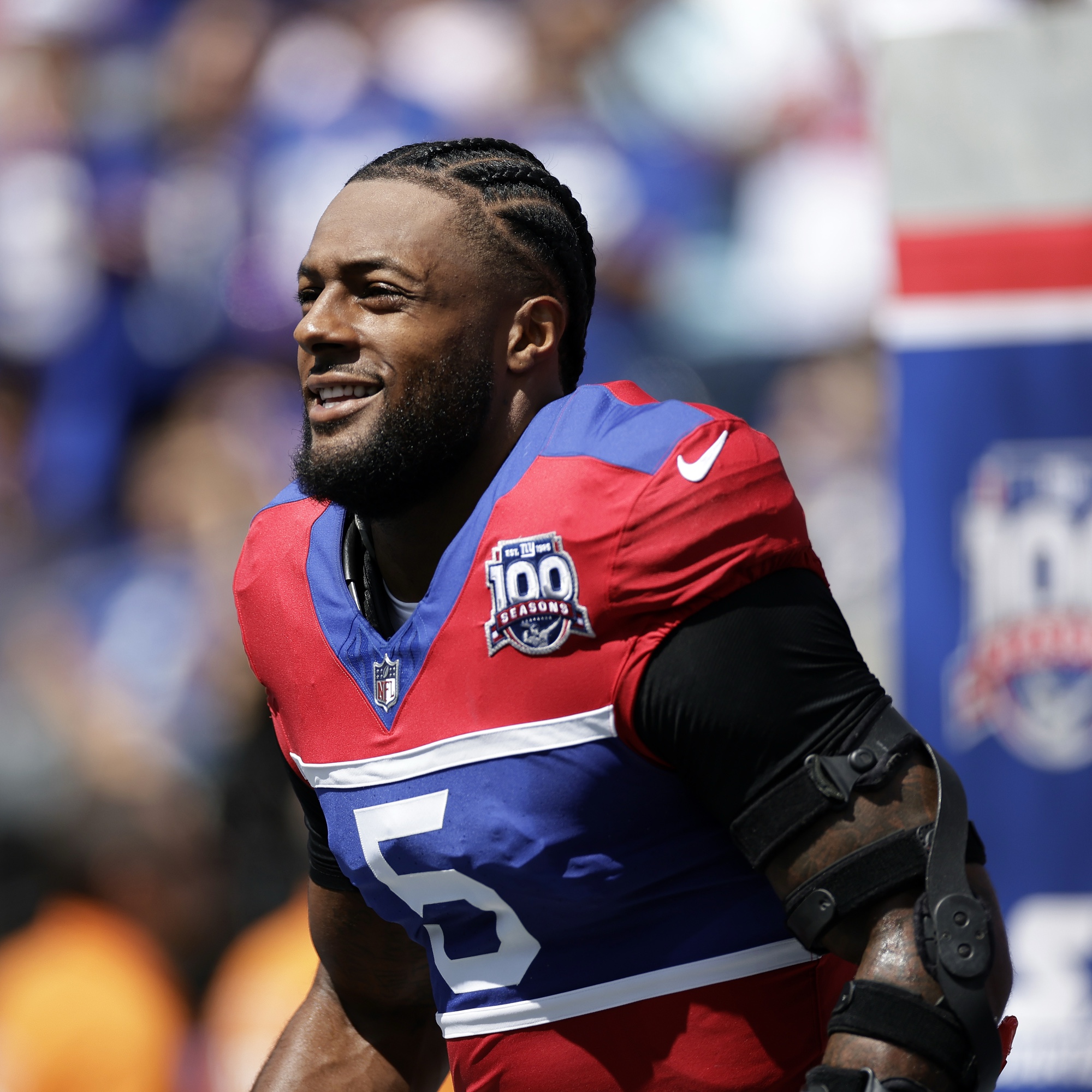 A football player in a red and blue uniform with the number 5, wearing a black arm brace, is on the field during a game.