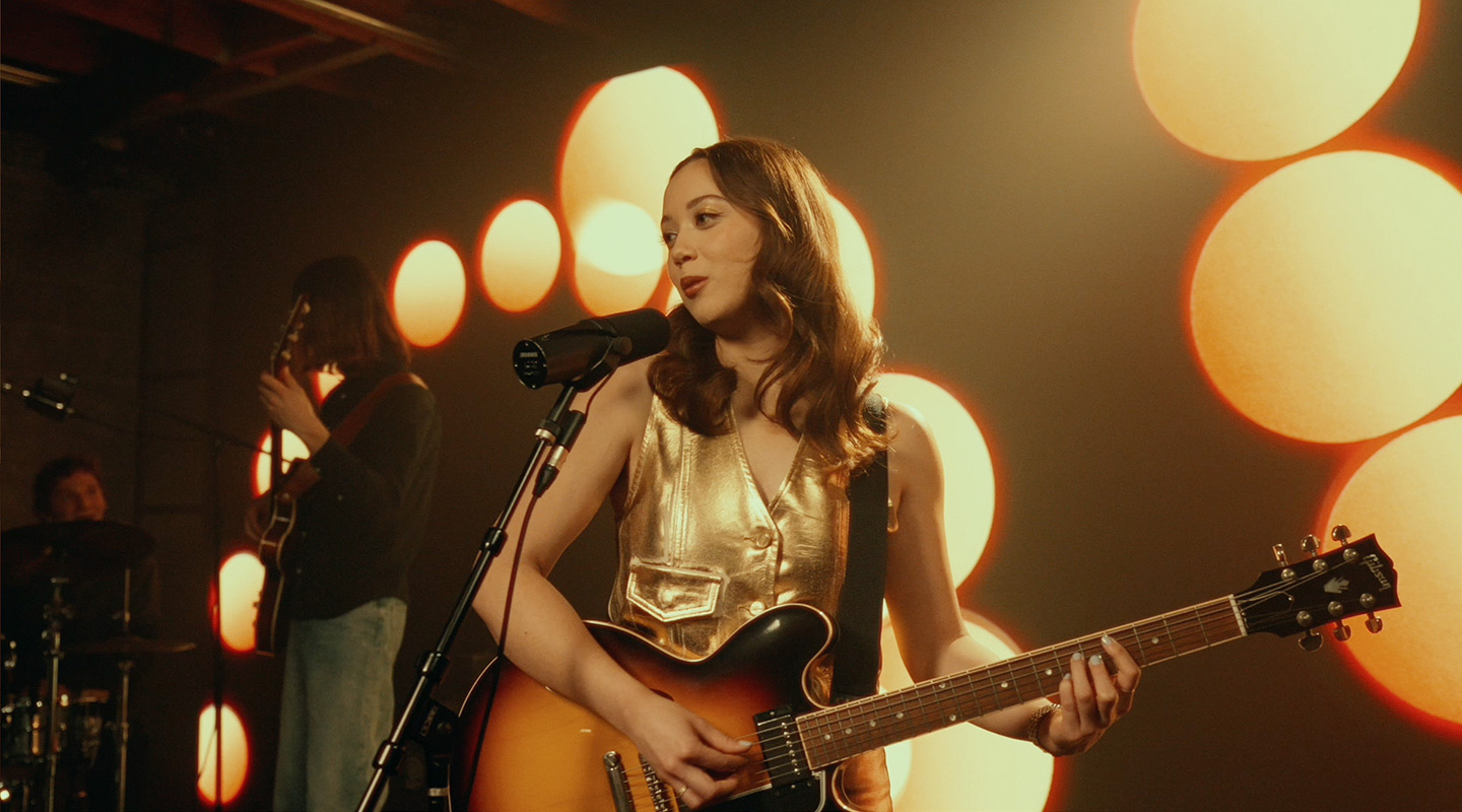 A female guitarist sings into a microphone on stage, flanked by a drummer and another guitarist, with large glowing orbs in the background.