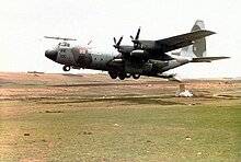 A Royal Air Force C-130 Hercules military plane. Its propellers attach to its wings, and it appears to be hovering just above a barren field. From the back of the aircraft, a package is being dropped onto the field from an open rear gangway.