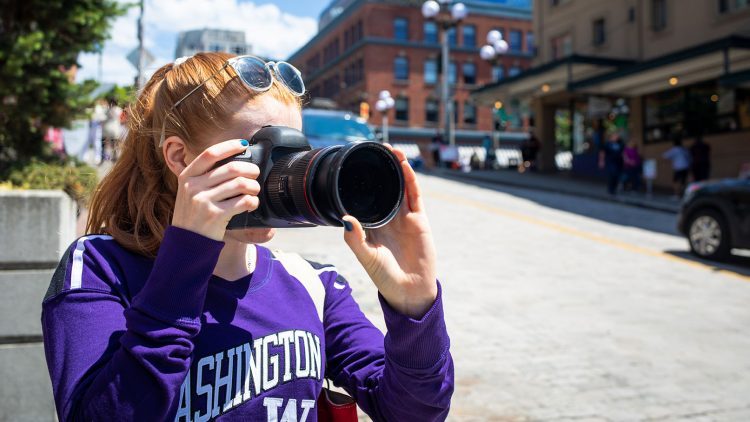 A woman in a purple UW shirt takes a picture on a downtown street corner.