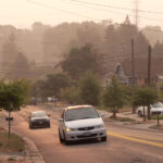 Photo of a neighborhood street with a couple cars driving on it. The air is orange and hazy.