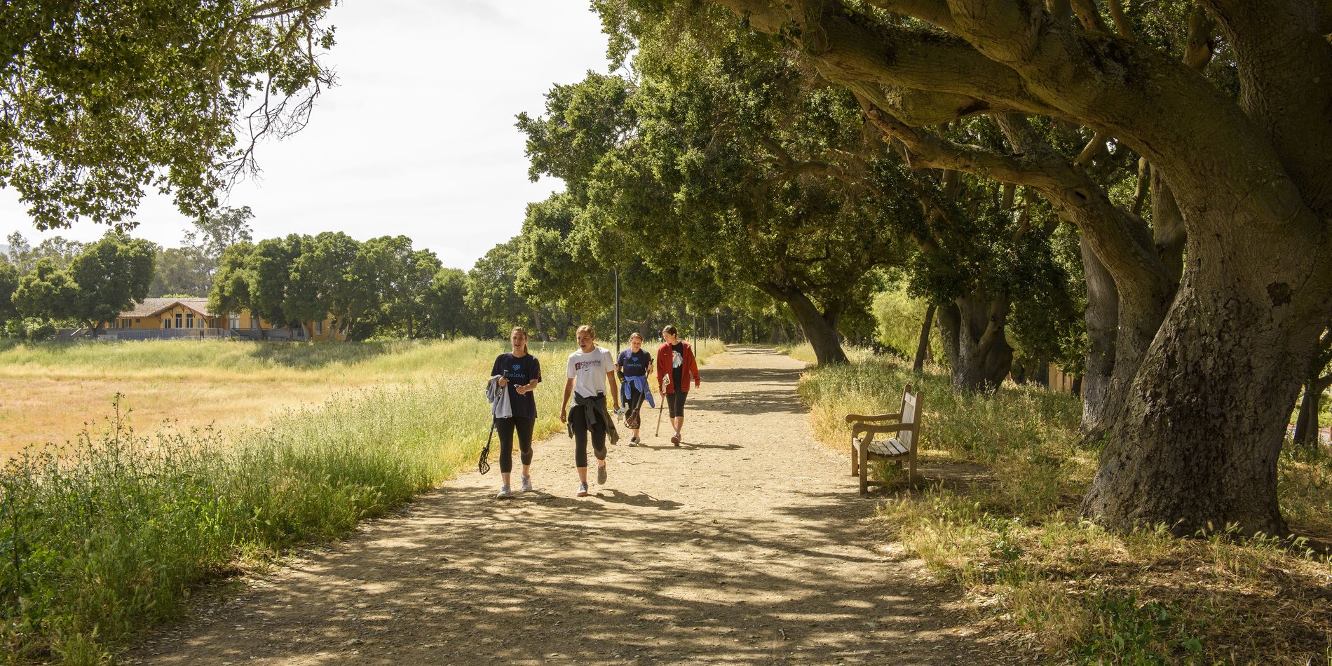 Small group of people walk along a tree lined path next to a dry Lake Laguinta on the Stanford Campus.