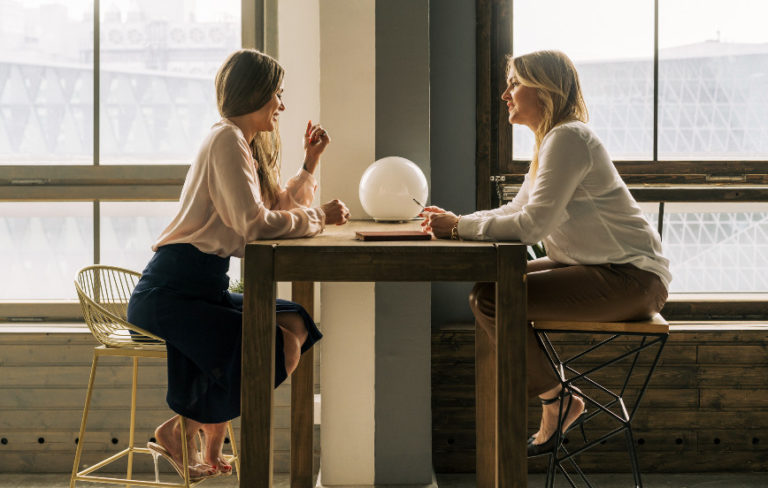 Two professional women with take away coffee cups sitting across from each other on tall stools at a tall wooden table