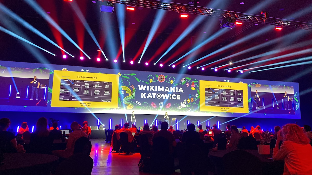 The main hall at Wikimania 2024 with a large screen showing Wikimania Katowice beneath lights shining out across the room, a crowd of people are sat in rows chairs as an audience facing the stage