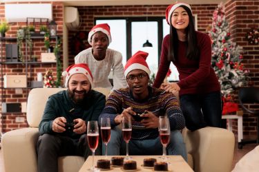 Young coworkers celebrating christmas in office, sitting on sofa and playing videogame with gamepads. Diverse workers team relaxing and having fun at new year holiday party in decorated office