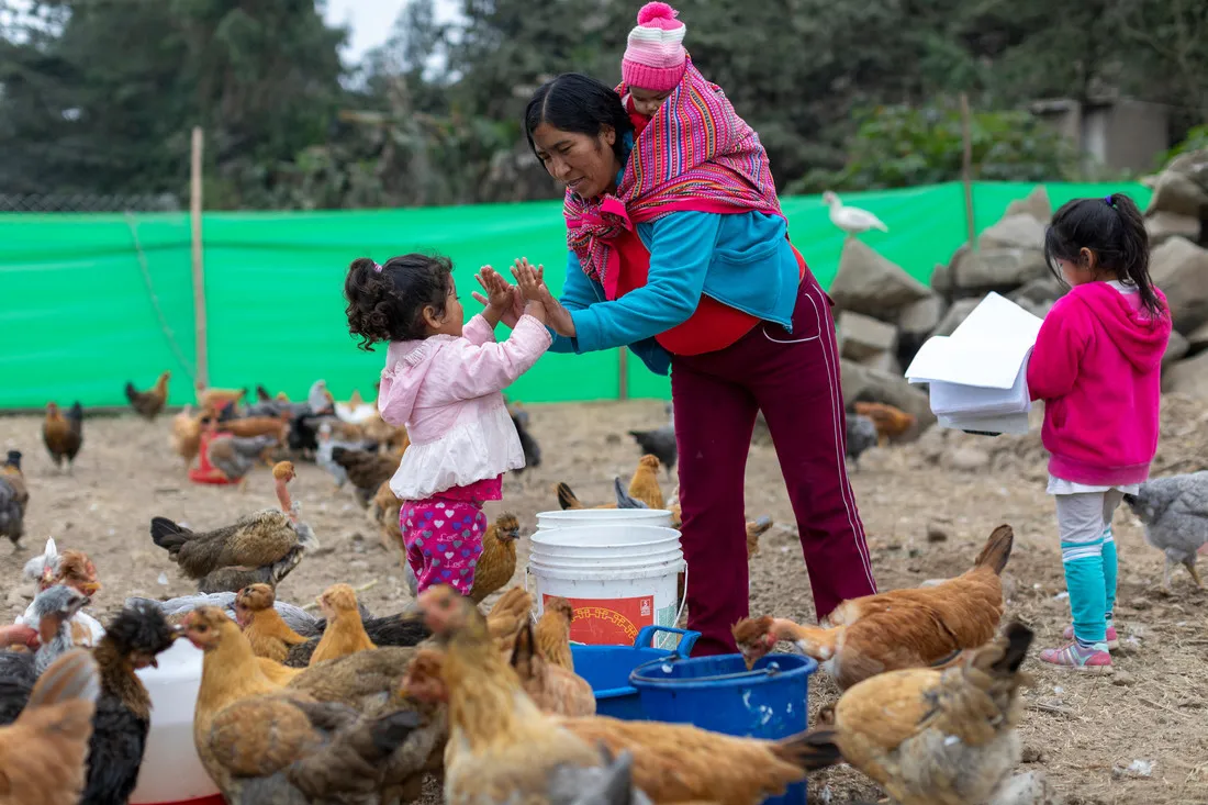 A woman carrying a baby on her shoulders high fives a young girl with both hands. They're surrounded by chickens eating on a farm.