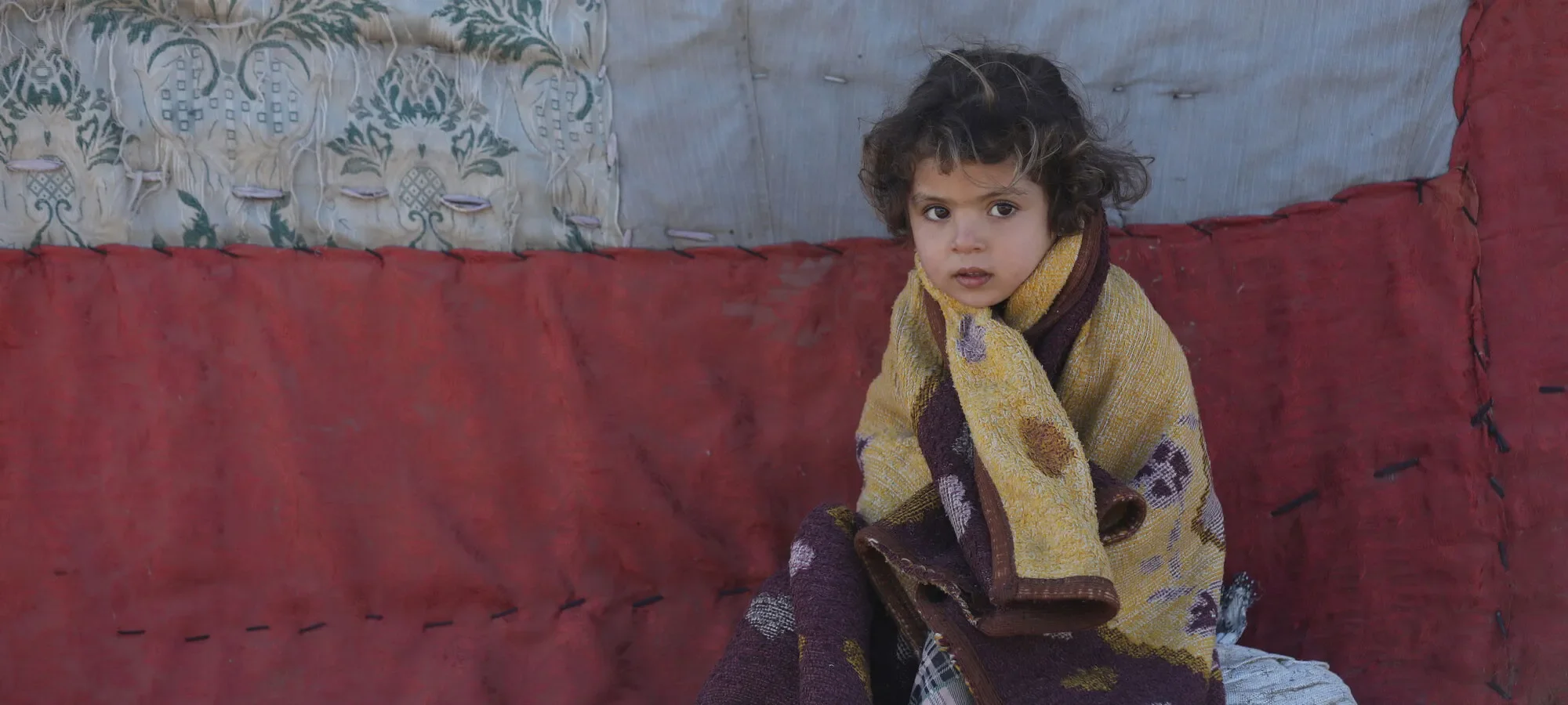 A young girl huddles outside a tent in a fleece blanket.