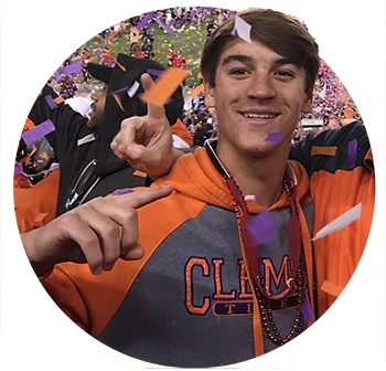 A male student and a man both wearing orange Clemson gear in the stands of a football stadium as orange and purple confetti rains down. 