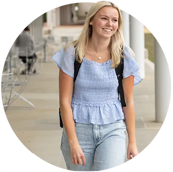 A female student wearing light-wash jeans and a light blue short-sleeved blouse carries a black backpack and walks across campus.