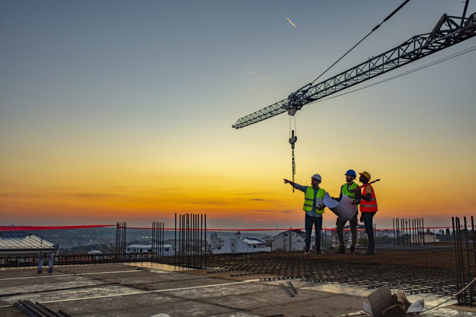 Three construction workers in uniform standing at construction site with crane in background, discussing building plans while holding blueprint at sunset under the tower crane.