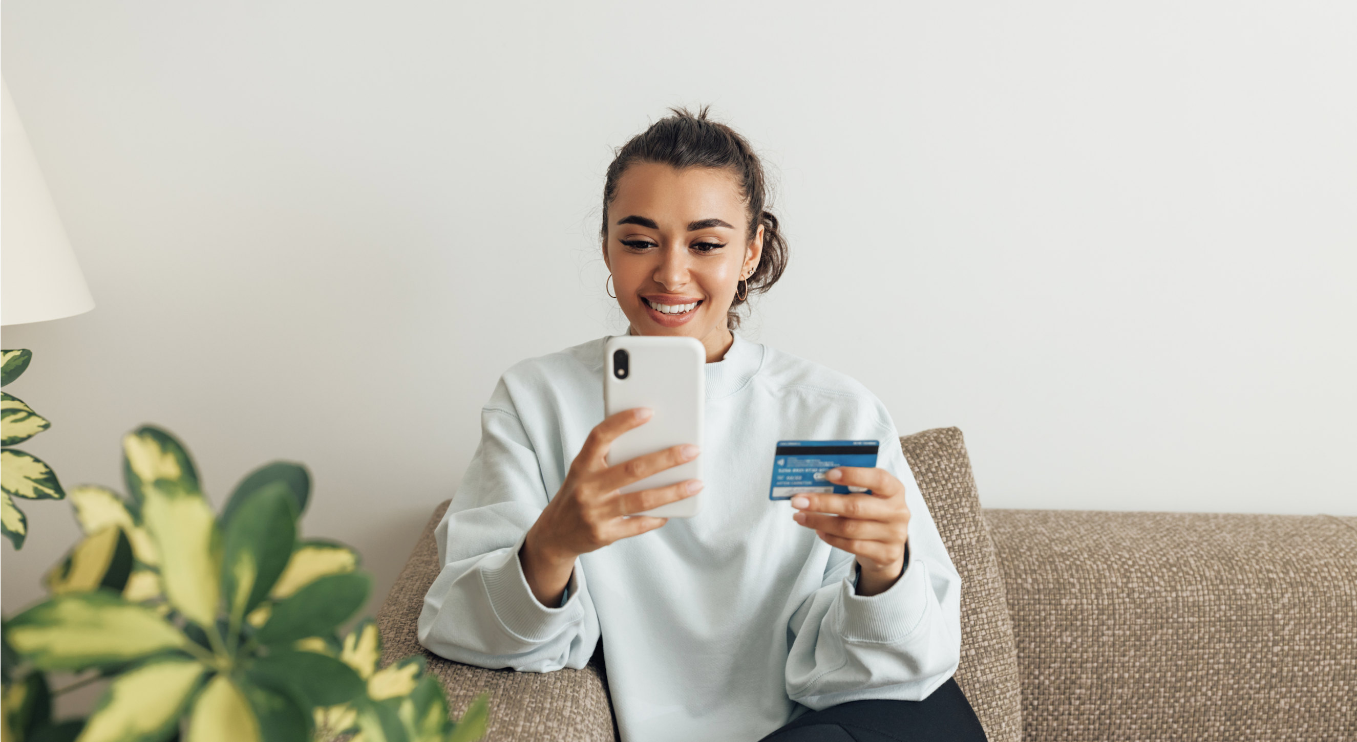 A young woman in a light blue top uses a credit card to complete a payment on her smartphone.