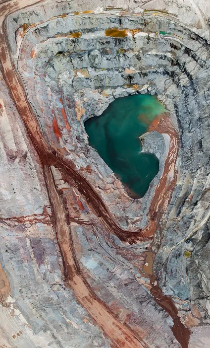 Aerial view of a mining site with distinct layers of red, orange, and gray earth surrounding a vibrant green water pool. The image showcases the vast scale and intricate patterns of the mining landscape, representing the transformative impact of infrastructure projects like the Simandou mining venture in Africa