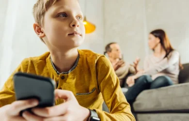 A child uses their smartphone while their parents talk on the sofa in the background.