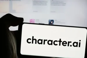 A hand holds a smartphone that reads character.ai in front of a computer screen.