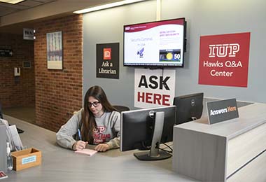 Student writing on a notepad at the LIbrary Information Desk. Signs read Ask a Librarian, Hawks Q&A Center, and Ask Here. 