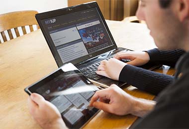 two students sitting at a desk, one using a tablet, the other a computer