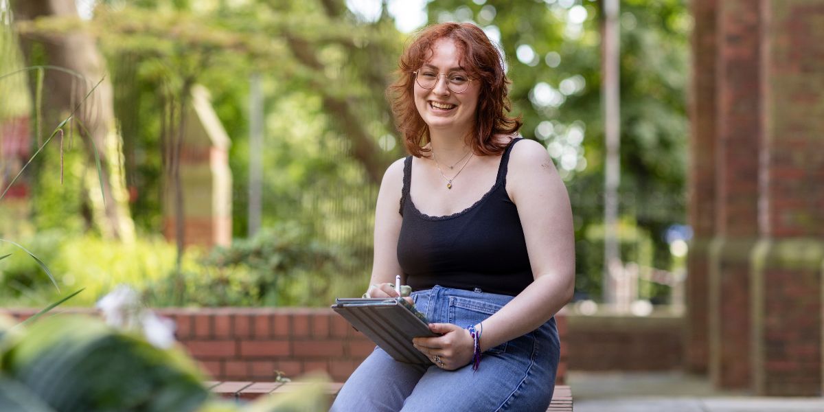 A student sat on a wall in a leafy street, using a tablet computer. They are smiling at the camera.