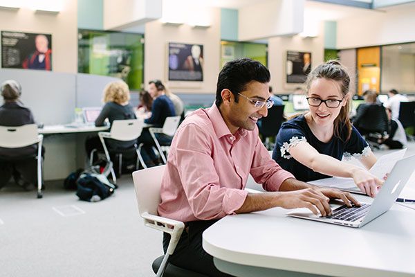 Two masters students in the library discussing work around a laptop