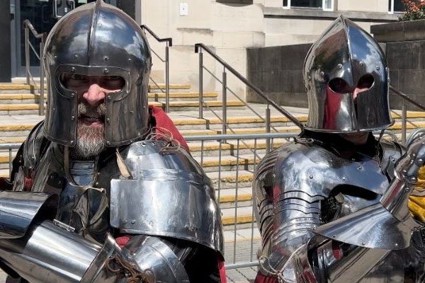 Two people dressed in armour pose for the camera, holding their swords up,