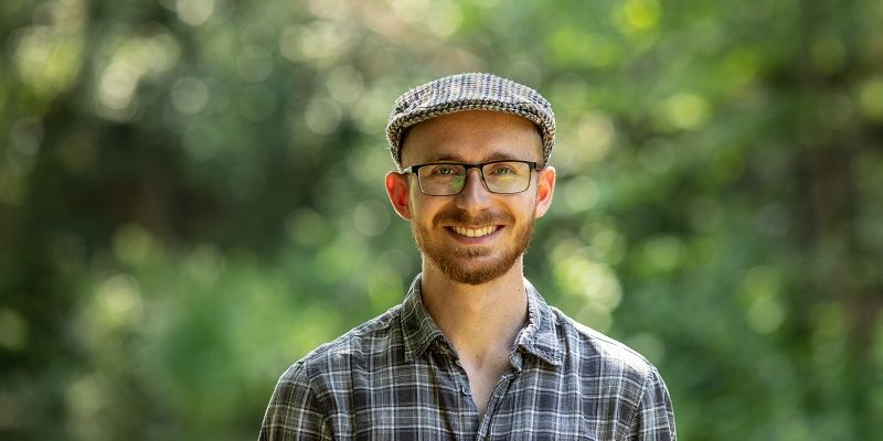 A student smiling at the camera with dappled light and trees in the background