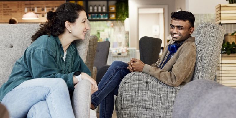 Two students sat in a cafe on sofas, chatting and smiling