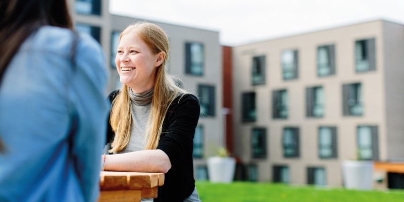 A smiling student sits at a picnic table in front of St Marks Residence talking to others.