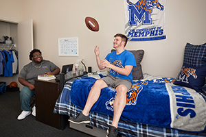 two male students in dorm room, 1 tossing football