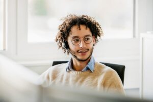 Office employee wearing a wireless headset, talks to clients while working at his computer in the office.