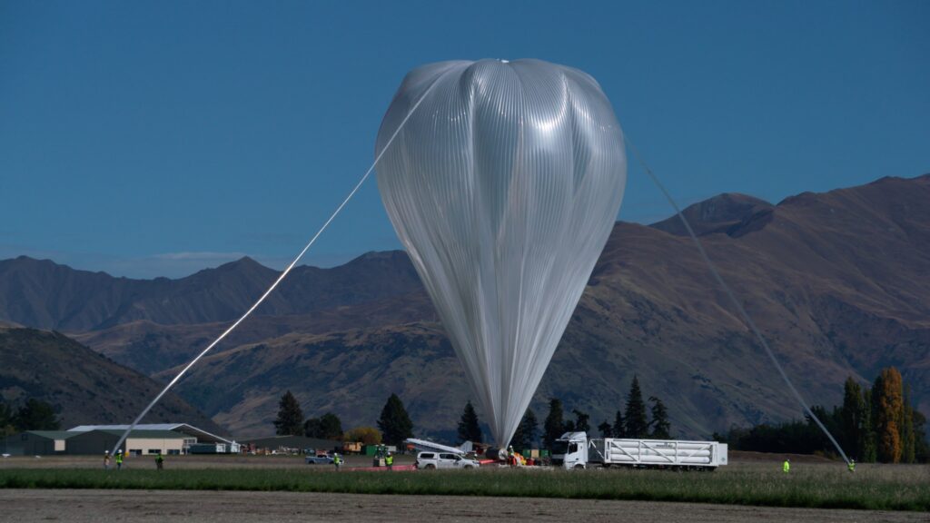 Large, translucent-white science balloon dominating a mountainous landscape