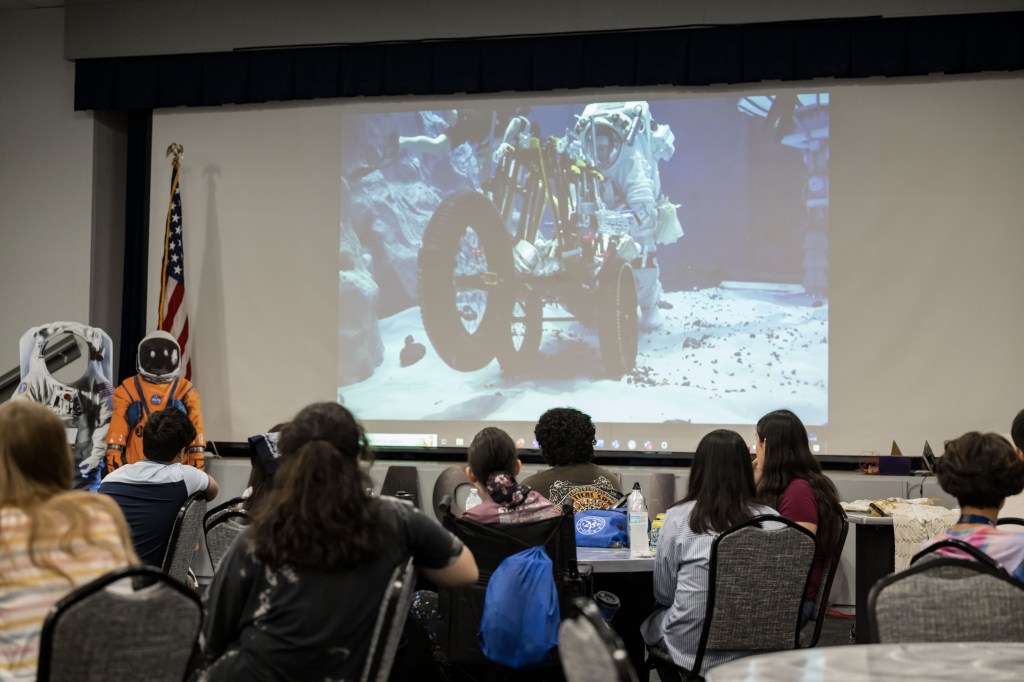 High school students sit with their backs to the camera as they watch a large screen displaying a white extravehicular activity suit being tested