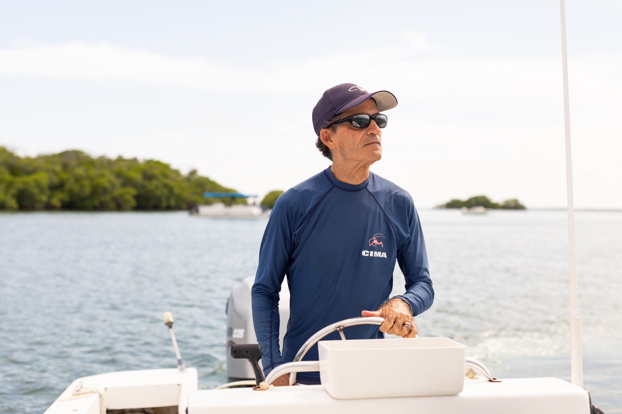 A Puerto Rican man wearing sunglasses, a navy baseball cap and longsleeve shirt is visible from the waist up, standing behind the silver wheel of a small white boat. In the background are a few islands of green foliage.