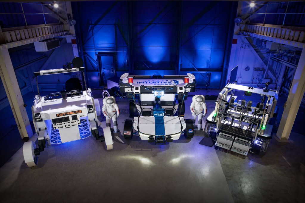 Three lunar terrain vehicles side by side inside of a large chamber with dramatic lighting. Astrolab's lunar terrain vehicle is white with the their orange logo on the front of the rover. Intuitive Machines' lunar terrain vehicle is white with a blue stripe on the front and their logo on the top of their rover. Lunar Outpost's lunar terrain vehicle is grey with green lights.