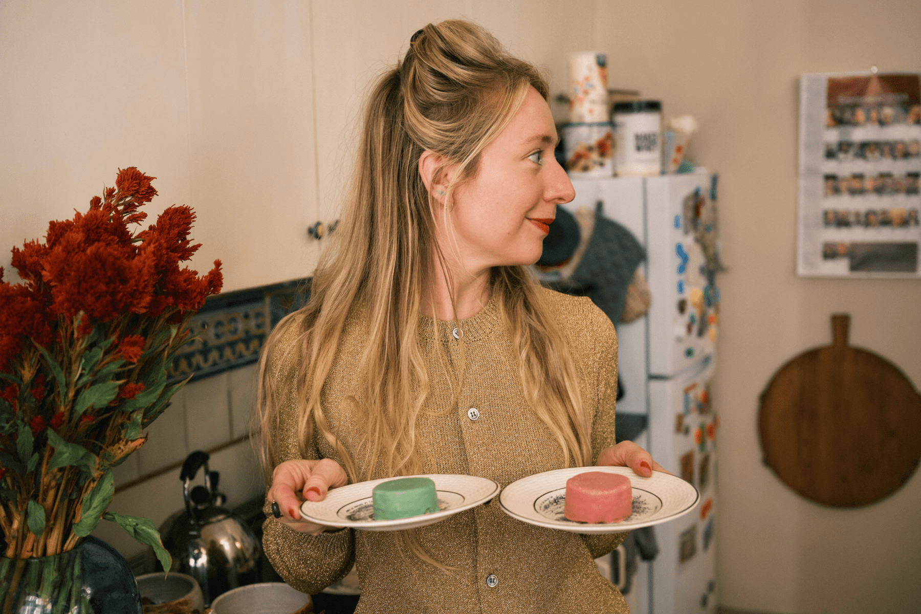 Joana Avillez in a gold sweater holds two dessert plates, one with a green cake and the other with a red cake.