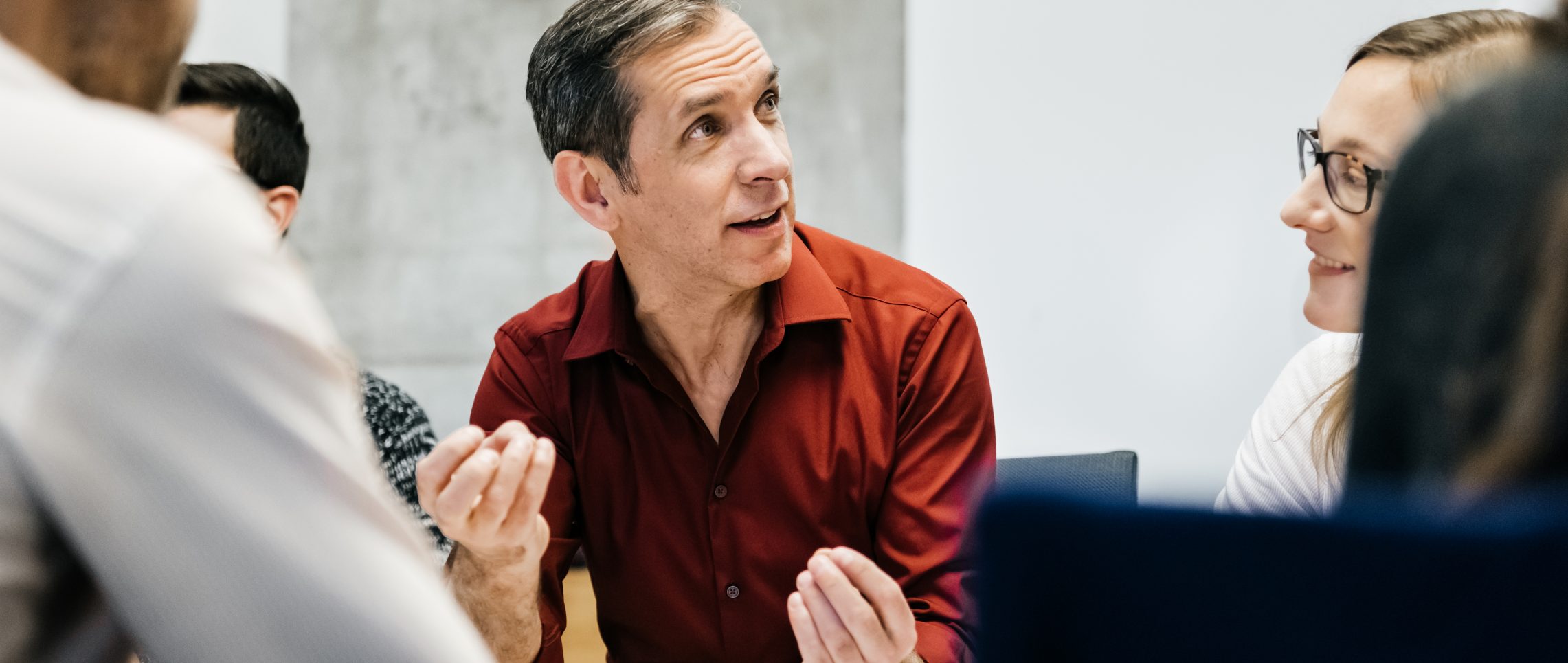 A caucasian man is speaking in a business meeting surrounded by colleagues listening to him. He uses his hands and seems convinced and focused.