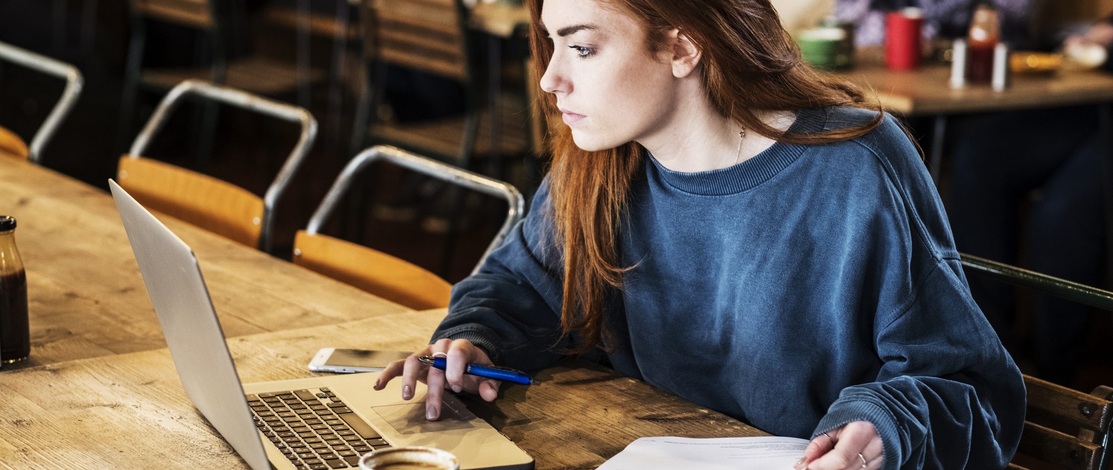 Young woman with long red hair sitting at table, working on laptop computer.