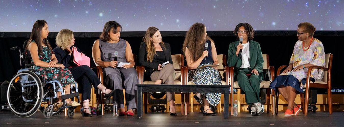 A panel of women of various backgrounds sits on stage with a background of stars behind them. One of the panelists is sitting in a wheelchair.