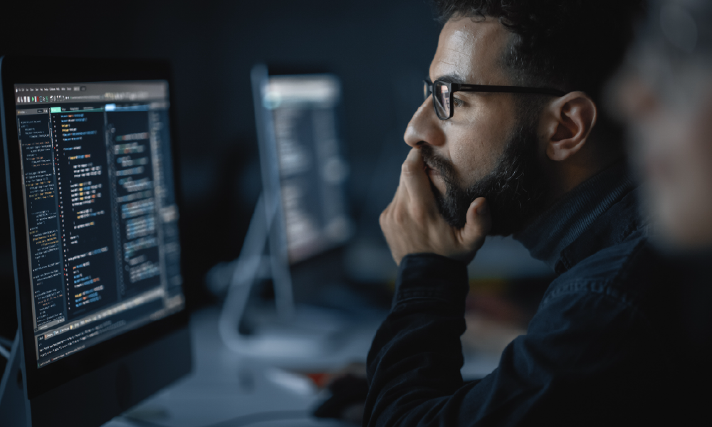 A man in black glasses and a sweater sits at a desktop monitor in a dimly lit room, deploying code. 