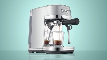 A Breville Bambino Plus espresso machine dispensing a shot of espresso into a clear glass, against a green background.